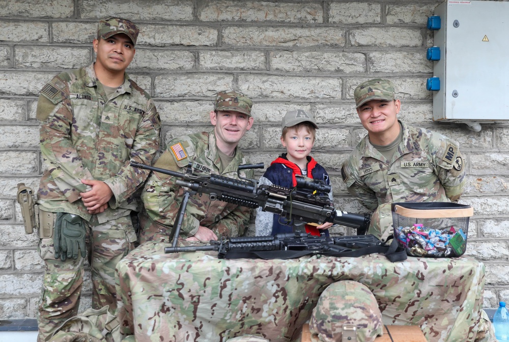 Three U.S. Army soldiers in MultiCam (OCP) uniforms pose with a young child at what appears to be a military open house or public outreach event. A display table covered with a camouflage cloth holds what appears to be a machine gun (likely an M240 or similar weapon) for public viewing. Name tapes visible include 'ALONSO', 'DUTCHER', and 'HTOO', with one soldier bearing an 'MP' (Military Police) patch.