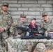 Three U.S. Army soldiers in MultiCam (OCP) uniforms pose with a young child at what appears to be a military open house or public outreach event. A display table covered with a camouflage cloth holds what appears to be a machine gun (likely an M240 or similar weapon) for public viewing. Name tapes visible include 'ALONSO', 'DUTCHER', and 'HTOO', with one soldier bearing an 'MP' (Military Police) patch.