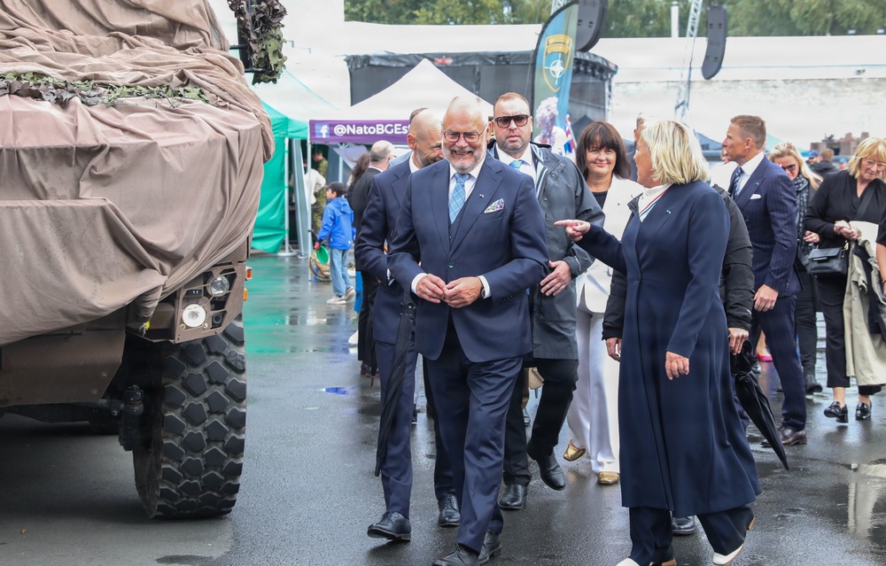 A group of formally dressed dignitaries or officials are walking through what appears to be a NATO military exhibition or defense expo, as indicated by the visible '@NatoBGEst' banner and NATO logo in the background. A smiling man in a navy suit with a light blue tie is prominently featured in the center, accompanied by a woman in a navy coat and other suited individuals. The wet pavement suggests rainy weather, and a camouflage-covered military vehicle is visible on the left.