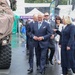 A group of formally dressed dignitaries or officials are walking through what appears to be a NATO military exhibition or defense expo, as indicated by the visible '@NatoBGEst' banner and NATO logo in the background. A smiling man in a navy suit with a light blue tie is prominently featured in the center, accompanied by a woman in a navy coat and other suited individuals. The wet pavement suggests rainy weather, and a camouflage-covered military vehicle is visible on the left.