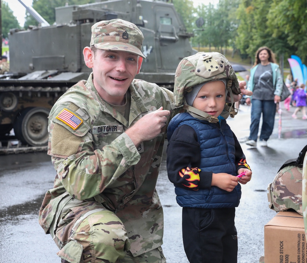A U.S. Army soldier named Dutcher, wearing OCP (Operational Camouflage Pattern) uniform, crouches beside a young child who is wearing an oversized military combat helmet. The scene appears to be a public military display or community outreach event, taking place outdoors in rainy conditions. A military armored vehicle is visible in the background along with civilian bystanders.