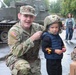 A U.S. Army soldier named Dutcher, wearing OCP (Operational Camouflage Pattern) uniform, crouches beside a young child who is wearing an oversized military combat helmet. The scene appears to be a public military display or community outreach event, taking place outdoors in rainy conditions. A military armored vehicle is visible in the background along with civilian bystanders.