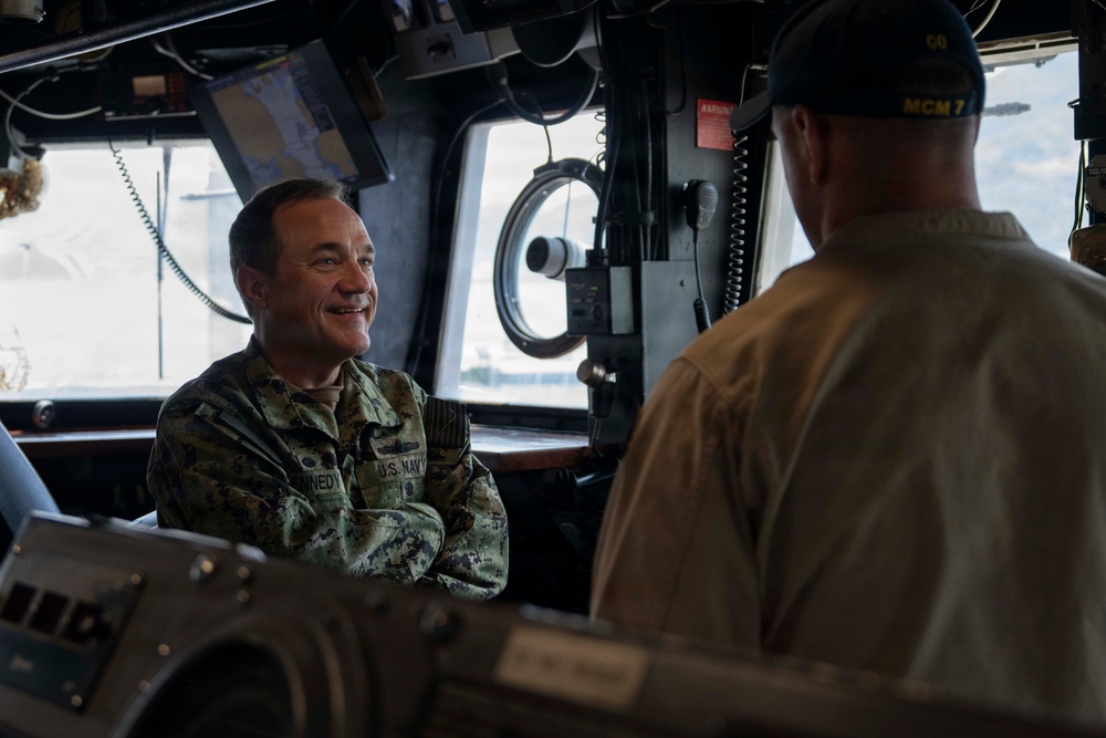 Two U.S. Navy personnel are conversing inside what appears to be the bridge or pilothouse of a naval vessel. The person facing the camera is wearing U.S. Navy camouflage (NWU) uniform with a name tape partially reading 'NEDY' and is smiling with arms crossed. The person with their back to the camera is wearing a tan uniform and a dark cap marked 'MCM 7' and 'CO', indicating they are the Commanding Officer of MCM-7 (a mine countermeasures ship).