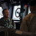 Two U.S. Navy personnel are conversing inside what appears to be the bridge or pilothouse of a naval vessel. The person facing the camera is wearing U.S. Navy camouflage (NWU) uniform with a name tape partially reading 'NEDY' and is smiling with arms crossed. The person with their back to the camera is wearing a tan uniform and a dark cap marked 'MCM 7' and 'CO', indicating they are the Commanding Officer of MCM-7 (a mine countermeasures ship).