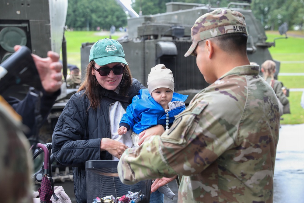 A U.S. Army soldier with the name tape 'ALONZO' interacts with a civilian woman holding an infant at what appears to be a military public outreach or open day event. The soldier is in OCP (Operational Camouflage Pattern) uniform and appears to be offering something from a container to the visitors. A military armored vehicle is visible in the background on a grassy area.