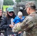 A U.S. Army soldier with the name tape 'ALONZO' interacts with a civilian woman holding an infant at what appears to be a military public outreach or open day event. The soldier is in OCP (Operational Camouflage Pattern) uniform and appears to be offering something from a container to the visitors. A military armored vehicle is visible in the background on a grassy area.