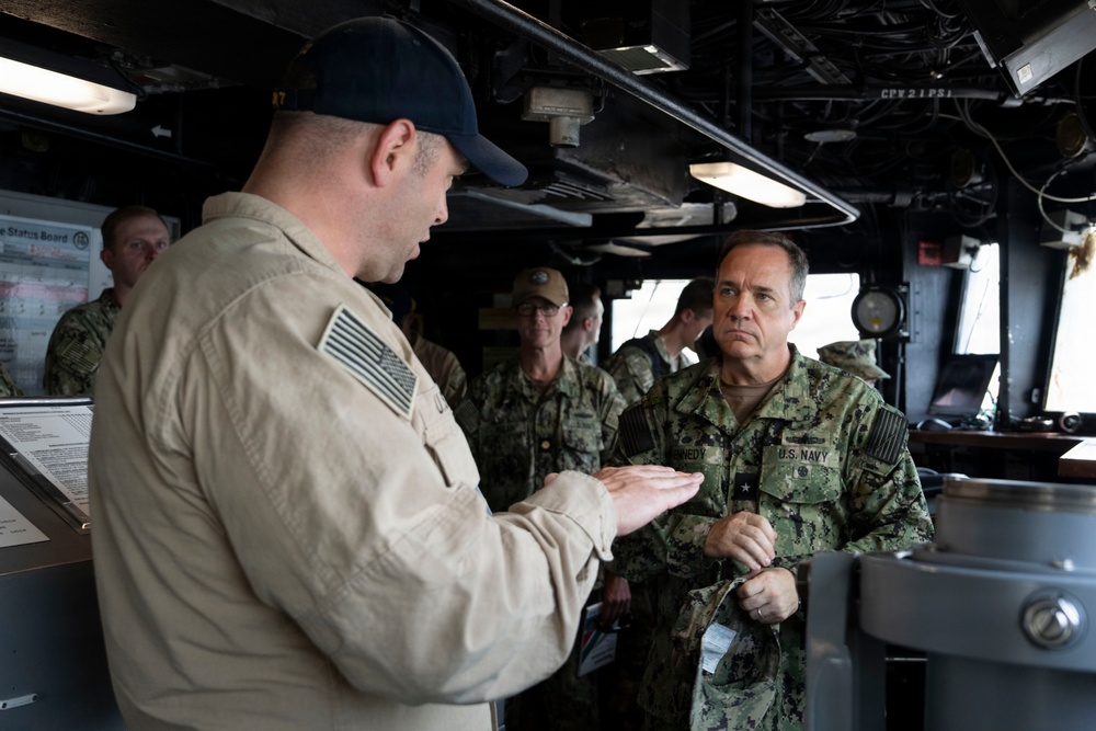 The image shows a briefing or discussion taking place on what appears to be the bridge or command area of a U.S. Navy vessel. A sailor in a tan/khaki flight suit is gesturing and speaking to a senior U.S. Navy officer in camouflage NWU (Navy Working Uniform) with visible rank insignia and a name tag partially reading 'NEDY'. Several other Navy personnel in NWU camouflage uniforms are present in the background.