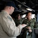 The image shows a briefing or discussion taking place on what appears to be the bridge or command area of a U.S. Navy vessel. A sailor in a tan/khaki flight suit is gesturing and speaking to a senior U.S. Navy officer in camouflage NWU (Navy Working Uniform) with visible rank insignia and a name tag partially reading 'NEDY'. Several other Navy personnel in NWU camouflage uniforms are present in the background.