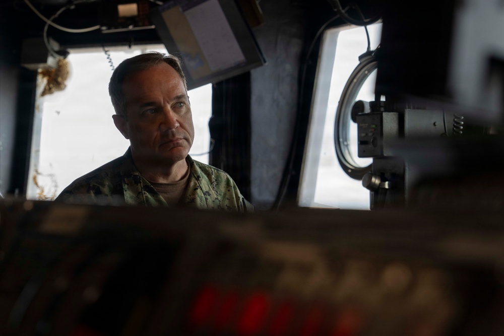 A male individual wearing military camouflage uniform is seated or positioned within what appears to be a naval vessel's bridge or command station. He is looking attentively to one side, surrounded by various electronic equipment, monitors, and navigation instruments. The lighting is dramatic with strong contrast between shadow and light.