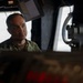 A male individual wearing military camouflage uniform is seated or positioned within what appears to be a naval vessel's bridge or command station. He is looking attentively to one side, surrounded by various electronic equipment, monitors, and navigation instruments. The lighting is dramatic with strong contrast between shadow and light.