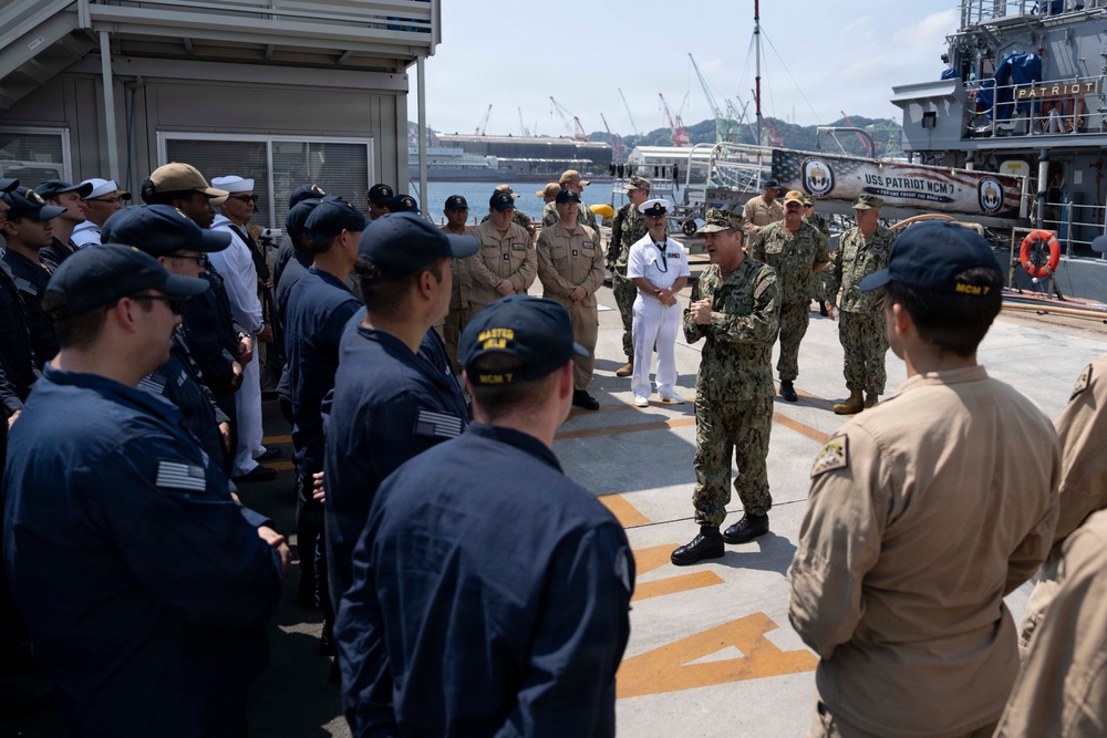 A military briefing or address is taking place on the deck of USS Patriot MCM 7, a U.S. Navy mine countermeasures ship. A senior officer in camouflage utilities is speaking to a formation of sailors wearing a mix of Navy Working Uniforms and tan/khaki uniforms. The setting appears to be a naval base with shipyard cranes and harbor infrastructure visible in the background.