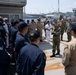 A military briefing or address is taking place on the deck of USS Patriot MCM 7, a U.S. Navy mine countermeasures ship. A senior officer in camouflage utilities is speaking to a formation of sailors wearing a mix of Navy Working Uniforms and tan/khaki uniforms. The setting appears to be a naval base with shipyard cranes and harbor infrastructure visible in the background.