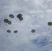 The image shows a large-scale military airdrop operation with numerous parachutes deployed across an overcast sky. Multiple cargo parachutes in olive/gray coloring are visible at various altitudes, with suspended loads beneath each canopy. In the background, additional parachutes and what appear to be recently deployed chutes are visible at higher altitude.