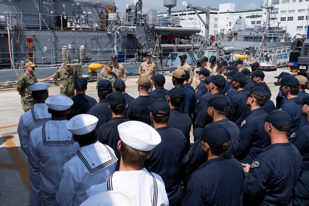 A group of U.S. Navy sailors assembled on the deck of a naval vessel at a port or naval base, appearing to receive a briefing or address from officers. The personnel are wearing a mix of dress whites, Navy Working Uniforms (NWU), and khaki uniforms, indicating a mix of enlisted and officer ranks. Multiple naval vessels and port infrastructure are visible in the background.