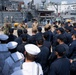 A group of U.S. Navy sailors assembled on the deck of a naval vessel at a port or naval base, appearing to receive a briefing or address from officers. The personnel are wearing a mix of dress whites, Navy Working Uniforms (NWU), and khaki uniforms, indicating a mix of enlisted and officer ranks. Multiple naval vessels and port infrastructure are visible in the background.