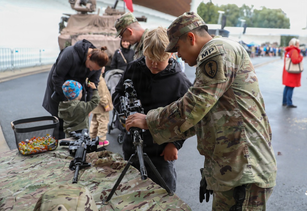A military open day or public exhibition event where uniformed soldiers are demonstrating weapons and equipment to civilians, including children. An MP (Military Police) soldier wearing OCP/MultiCam camouflage is showing a bipod-mounted rifle to a young boy at a display table. Additional weapons, equipment, and a military vehicle are visible in the background.