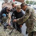 A military open day or public exhibition event where uniformed soldiers are demonstrating weapons and equipment to civilians, including children. An MP (Military Police) soldier wearing OCP/MultiCam camouflage is showing a bipod-mounted rifle to a young boy at a display table. Additional weapons, equipment, and a military vehicle are visible in the background.