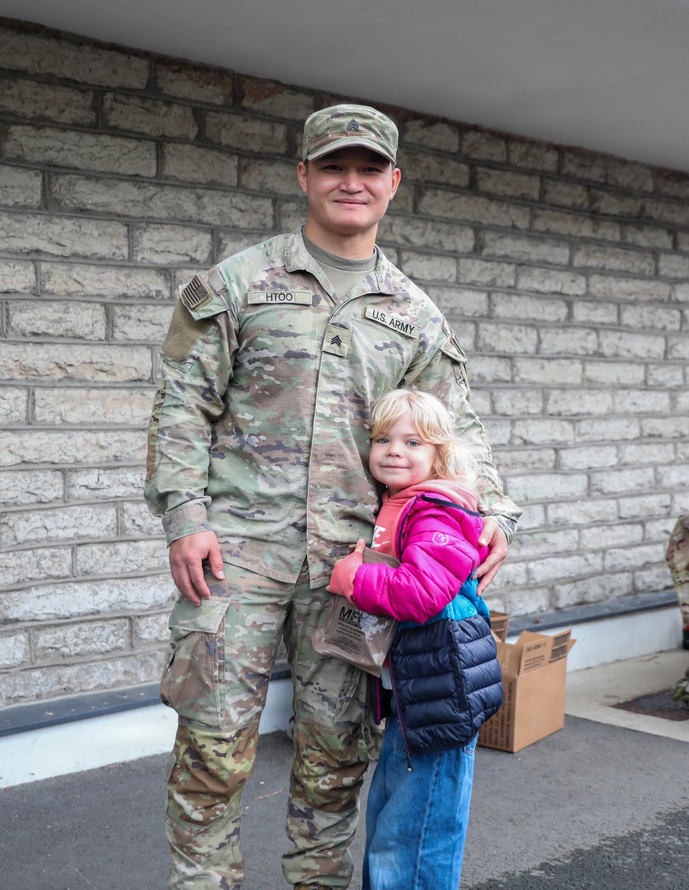 A U.S. Army Sergeant named Htoo, wearing OCP (Operational Camouflage Pattern) uniform, poses with a young blonde child who is hugging his side. The child is wearing a pink and navy puffer jacket and jeans, and appears to be smiling at the camera. They are standing in front of a light-colored brick wall, with a cardboard box visible in the background.