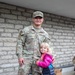 A U.S. Army Sergeant named Htoo, wearing OCP (Operational Camouflage Pattern) uniform, poses with a young blonde child who is hugging his side. The child is wearing a pink and navy puffer jacket and jeans, and appears to be smiling at the camera. They are standing in front of a light-colored brick wall, with a cardboard box visible in the background.