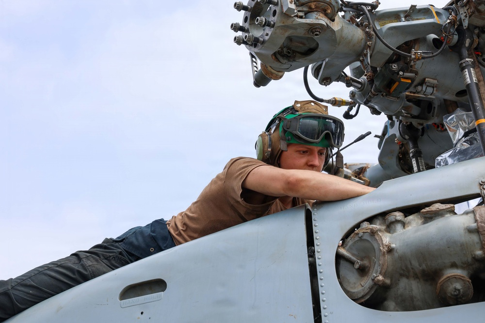 A military aviation mechanic or technician is performing maintenance on a helicopter, leaning over the fuselage near the rotor head assembly. The individual is wearing standard flight deck gear including a helmet with goggles and hearing protection. The image is taken from a low angle, emphasizing the complex mechanical components of the rotor system above.