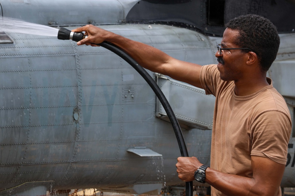 A man in a tan/brown t-shirt is using a hose with a spray nozzle to wash the exterior of a military aircraft. The aircraft appears to be a gray Navy plane with visible riveted panels and partial 'NAVY' lettering on the fuselage. The scene appears to be routine aircraft maintenance or cleaning.