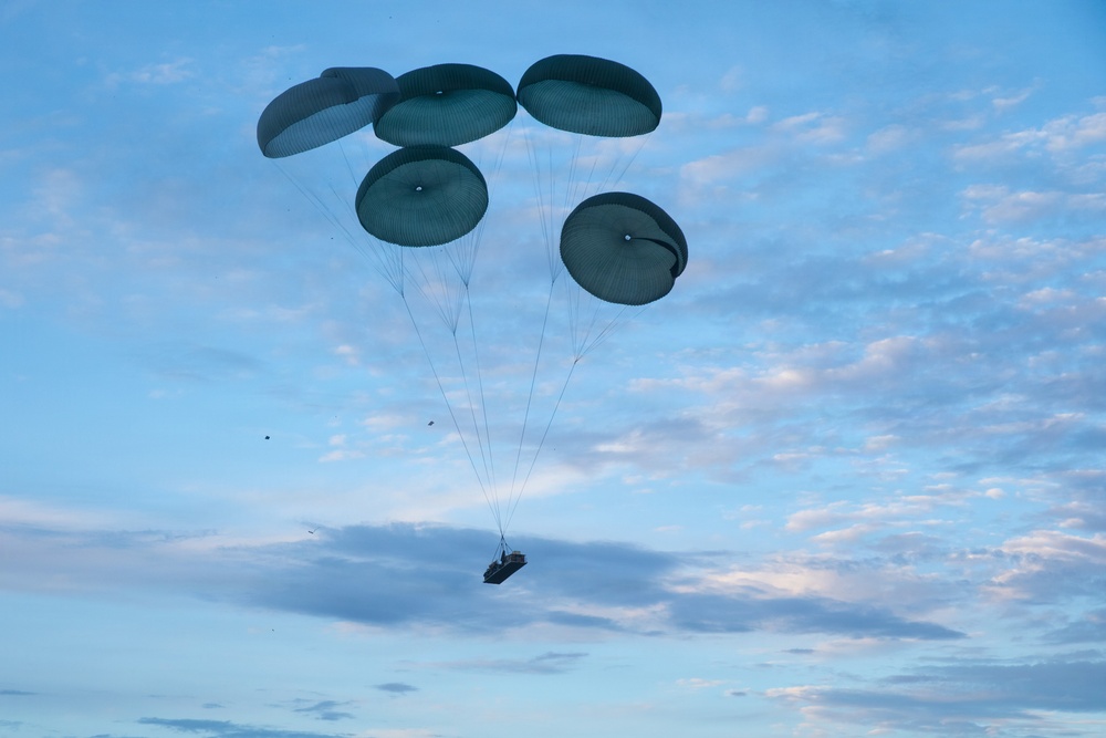 The image shows a military aerial delivery operation with multiple parachutes deployed to slow the descent of a cargo load. Five dark olive/green round parachutes are clustered together, suspending what appears to be a palletized cargo bundle beneath them. The scene is set against a partly cloudy blue sky, suggesting daytime conditions.