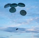 The image shows a military aerial delivery operation with multiple parachutes deployed to slow the descent of a cargo load. Five dark olive/green round parachutes are clustered together, suspending what appears to be a palletized cargo bundle beneath them. The scene is set against a partly cloudy blue sky, suggesting daytime conditions.