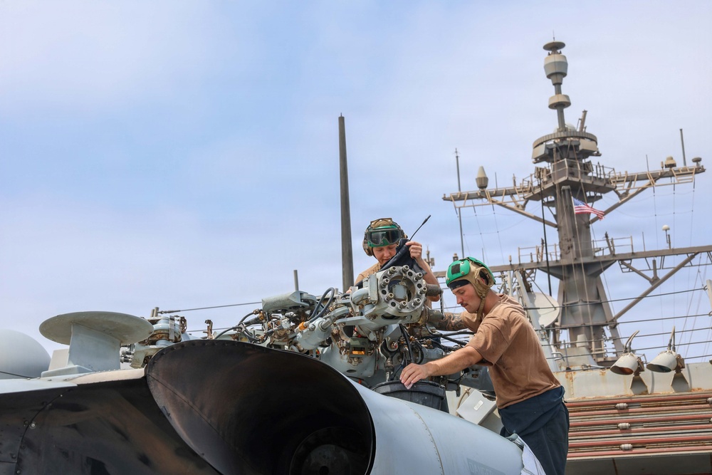 Two military personnel are performing maintenance on what appears to be a helicopter engine or rotor assembly on the deck of a U.S. Navy warship. Both individuals are wearing hearing protection and goggles, suggesting an active flight deck environment. A naval vessel's superstructure with radar arrays and an American flag is visible in the background.