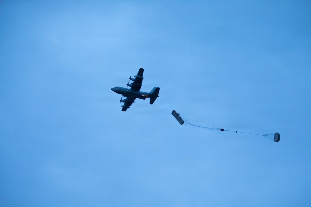 A military transport aircraft, consistent with a C-130 Hercules, is captured mid-flight against a blue sky while performing an aerial cargo drop. A pallet or container has been deployed from the aircraft's rear and is being slowed by an extraction/drogue parachute still in the process of deploying. The aircraft and payload are silhouetted, suggesting the photo was taken from below at an angle.