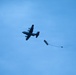 A military transport aircraft, consistent with a C-130 Hercules, is captured mid-flight against a blue sky while performing an aerial cargo drop. A pallet or container has been deployed from the aircraft's rear and is being slowed by an extraction/drogue parachute still in the process of deploying. The aircraft and payload are silhouetted, suggesting the photo was taken from below at an angle.