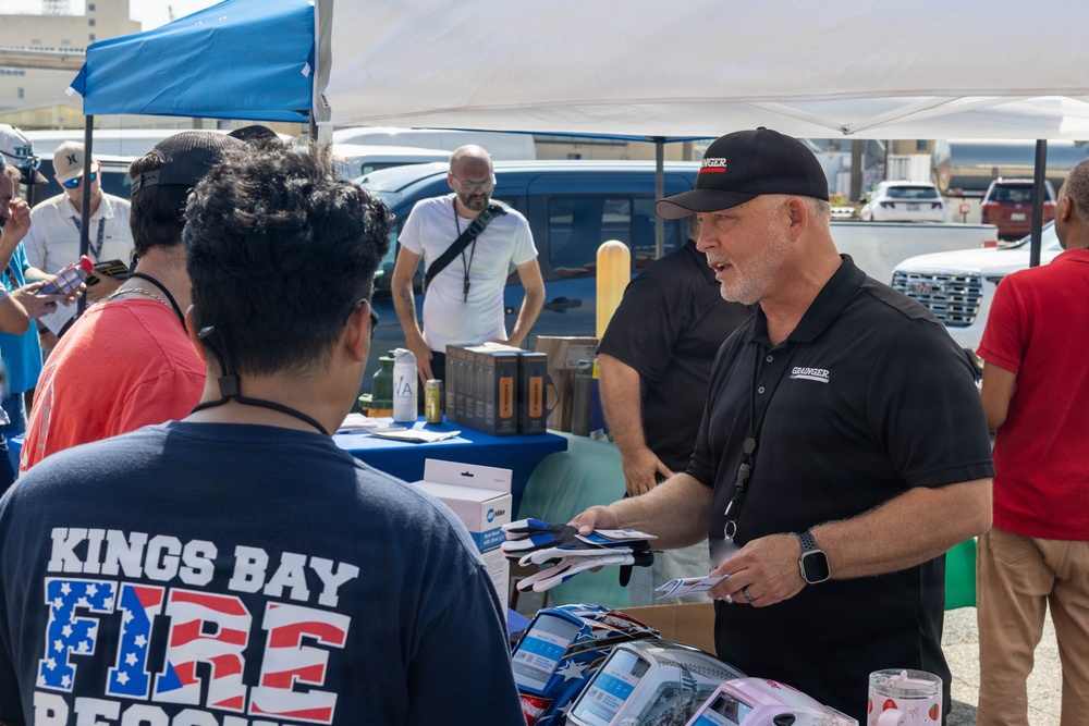 An outdoor vendor or trade event where a Grainger representative in a black polo shirt and cap is distributing materials, likely gloves or promotional items, to attendees. A person wearing a 'Kings Bay Fire Rescue' shirt is at the table receiving items. Multiple vendor booths with canopy tents are visible in the background.