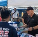 An outdoor vendor or trade event where a Grainger representative in a black polo shirt and cap is distributing materials, likely gloves or promotional items, to attendees. A person wearing a 'Kings Bay Fire Rescue' shirt is at the table receiving items. Multiple vendor booths with canopy tents are visible in the background.