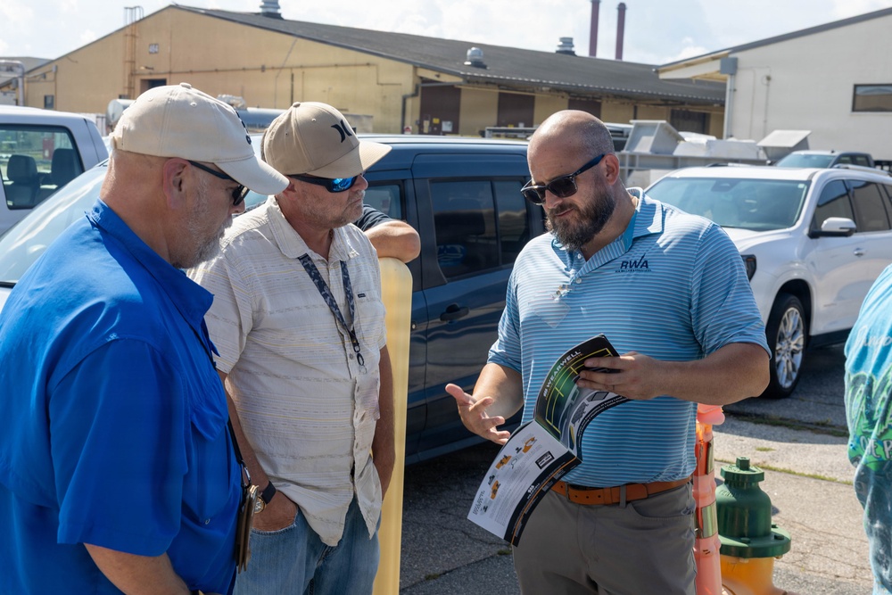 Three men are engaged in a professional discussion in an outdoor parking lot or industrial facility area. The man on the right, wearing an 'RWA' branded polo shirt, is presenting or explaining information from a printed brochure or catalog to the other two men. The setting appears to be a field meeting or site visit at an industrial or municipal facility.
