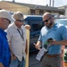 Three men are engaged in a professional discussion in an outdoor parking lot or industrial facility area. The man on the right, wearing an 'RWA' branded polo shirt, is presenting or explaining information from a printed brochure or catalog to the other two men. The setting appears to be a field meeting or site visit at an industrial or municipal facility.
