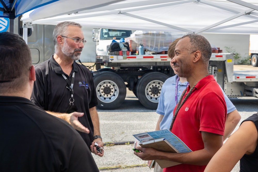 A group of people are engaged in conversation under a white canopy tent in an outdoor setting, likely at a work site or event. The man in the red polo shirt is holding a clipboard with visible documents, and the man in the black shirt is wearing a lanyard with credentials. Large flatbed trucks are visible in the background.