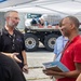 A group of people are engaged in conversation under a white canopy tent in an outdoor setting, likely at a work site or event. The man in the red polo shirt is holding a clipboard with visible documents, and the man in the black shirt is wearing a lanyard with credentials. Large flatbed trucks are visible in the background.