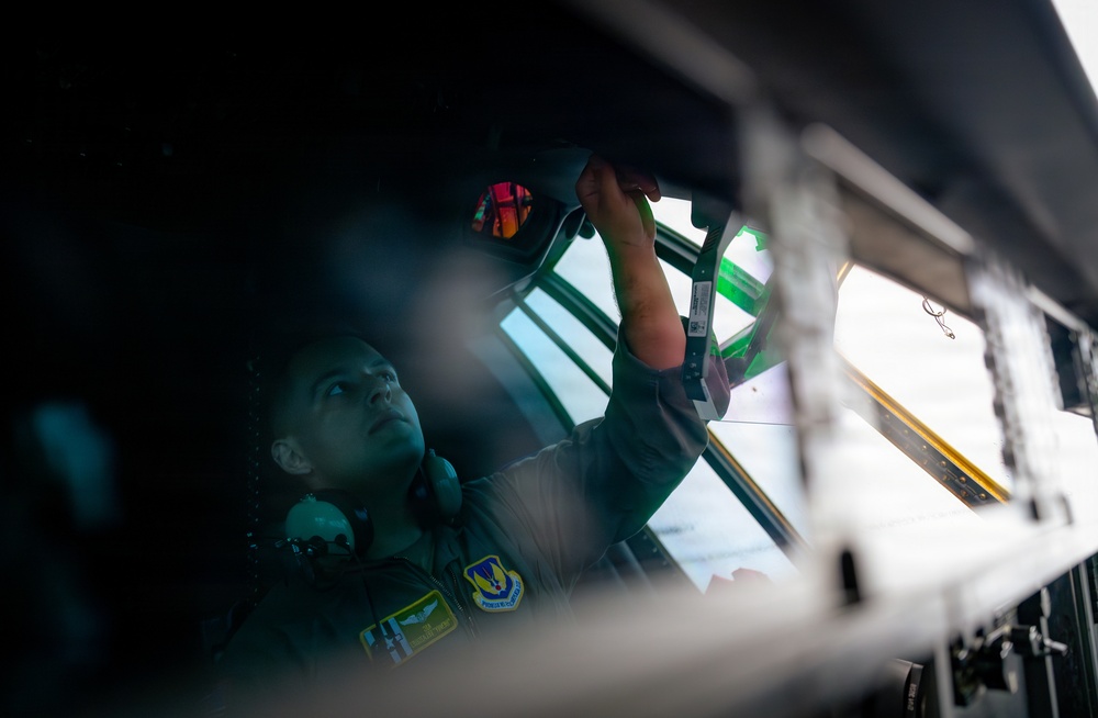 A military aviator in flight suit is working inside what appears to be a military aircraft cockpit or cargo aircraft interior. The individual is reaching upward to handle or inspect equipment mounted near the overhead area or window frame. The image is taken from a low angle with dramatic lighting, creating a moody, high-contrast composition.