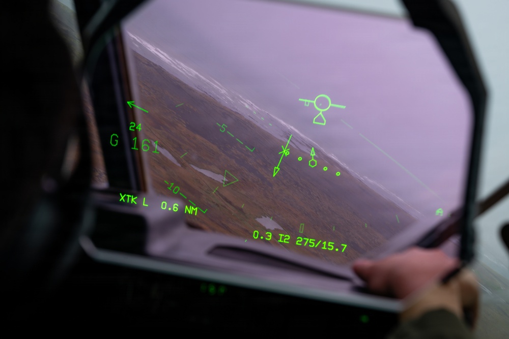 The image shows a view from inside a military aircraft cockpit, looking through the Head-Up Display (HUD) toward a terrain below. Green HUD symbology overlays the windscreen, displaying flight data including navigation, altitude, and targeting information. The aircraft appears to be in a banked or descending attitude over rugged, brownish terrain under overcast skies.