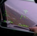 The image shows a view from inside a military aircraft cockpit, looking through the Head-Up Display (HUD) toward a terrain below. Green HUD symbology overlays the windscreen, displaying flight data including navigation, altitude, and targeting information. The aircraft appears to be in a banked or descending attitude over rugged, brownish terrain under overcast skies.
