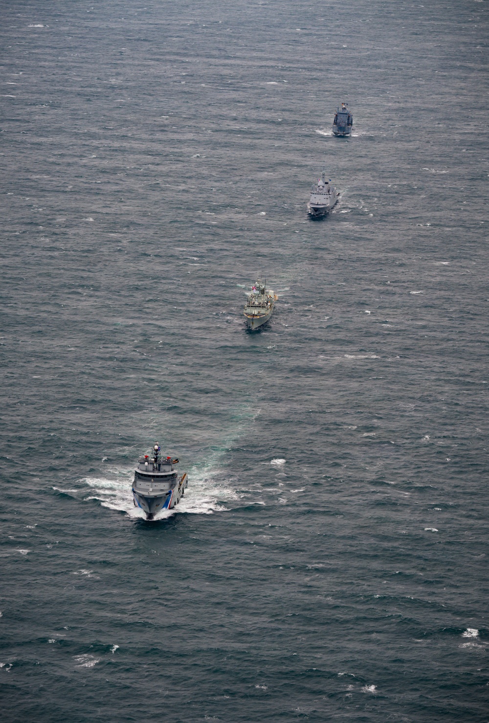 An aerial photograph shows four naval/coast guard vessels sailing in a single-file formation across open ocean. The ships appear to be traveling in the same direction, spaced evenly apart, suggesting a coordinated maritime exercise or transit operation. The lead vessel displays distinctive blue and white hull markings consistent with a coast guard or patrol vessel.