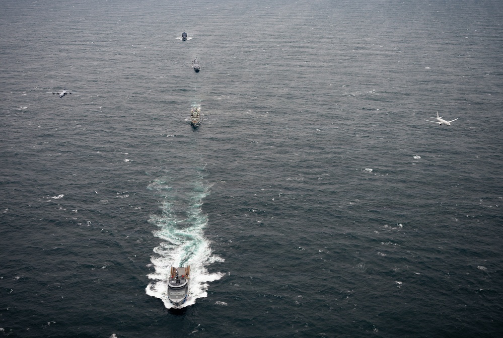 An aerial photograph captures a naval formation of multiple vessels traveling in a column across open ocean waters. Two aircraft are visible flanking the formation — one on the left appearing to be a military turboprop (possibly a C-130 Hercules) and one on the right appearing to be a maritime patrol jet aircraft. The lead vessel in the foreground is generating a prominent white wake, suggesting the formation is underway at moderate speed.