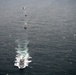 An aerial photograph captures a naval formation of multiple vessels traveling in a column across open ocean waters. Two aircraft are visible flanking the formation — one on the left appearing to be a military turboprop (possibly a C-130 Hercules) and one on the right appearing to be a maritime patrol jet aircraft. The lead vessel in the foreground is generating a prominent white wake, suggesting the formation is underway at moderate speed.