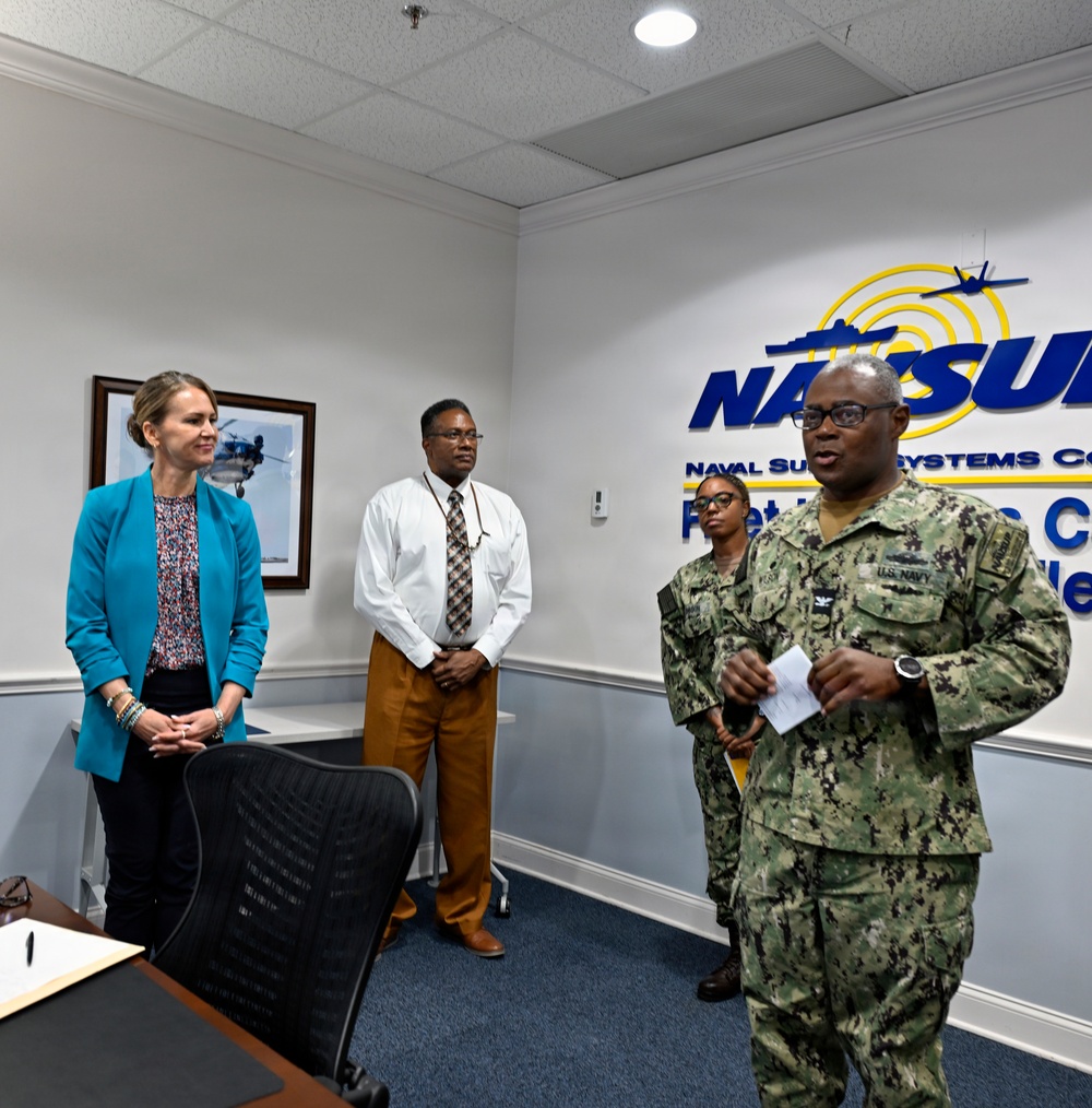 A formal meeting or briefing is taking place inside a Naval Supply Systems Command (NAVSUP) facility. A U.S. Navy officer in camouflage uniform appears to be addressing or presenting to a small group that includes a civilian woman in a teal blazer, a civilian man in business casual attire, and a junior Navy service member in the background. The NAVSUP logo and partial text are prominently displayed on the wall behind the group.