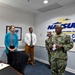A formal meeting or briefing is taking place inside a Naval Supply Systems Command (NAVSUP) facility. A U.S. Navy officer in camouflage uniform appears to be addressing or presenting to a small group that includes a civilian woman in a teal blazer, a civilian man in business casual attire, and a junior Navy service member in the background. The NAVSUP logo and partial text are prominently displayed on the wall behind the group.