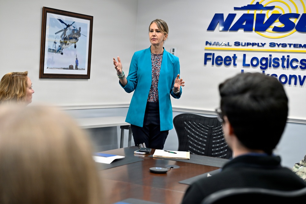 A woman in a teal blazer is presenting or speaking to a small group in a conference room at NAVSU (Naval Supply Systems) Fleet Logistics Jacksonville. She is standing at the head of a conference table with her hands raised in a gesturing motion. The setting appears to be a formal briefing or meeting environment within a U.S. Navy facility.