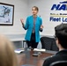 A woman in a teal blazer is presenting or speaking to a small group in a conference room at NAVSU (Naval Supply Systems) Fleet Logistics Jacksonville. She is standing at the head of a conference table with her hands raised in a gesturing motion. The setting appears to be a formal briefing or meeting environment within a U.S. Navy facility.