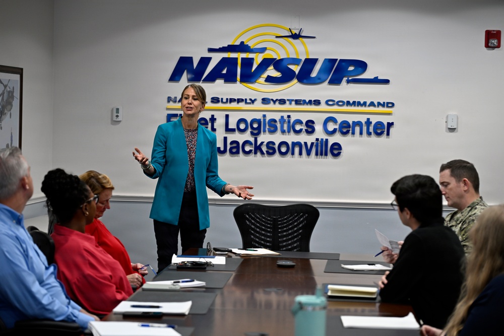 A woman in a teal blazer is presenting or briefing a small group of people seated around a conference table at NAVSUP Fleet Logistics Center Jacksonville. The setting is an official U.S. Navy Naval Supply Systems Command facility conference room. Attendees include both civilian personnel and at least one uniformed Navy sailor.