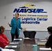 A woman in a teal blazer is presenting or briefing a small group of people seated around a conference table at NAVSUP Fleet Logistics Center Jacksonville. The setting is an official U.S. Navy Naval Supply Systems Command facility conference room. Attendees include both civilian personnel and at least one uniformed Navy sailor.