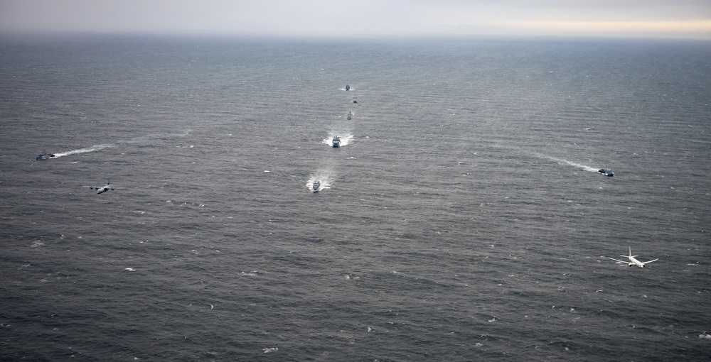 An aerial photograph shows a coordinated naval or maritime formation over open ocean under overcast skies. Multiple vessels are arranged in a column formation moving away from the camera, flanked by what appear to be aircraft flying at low altitude on either side. The scene suggests a military or coast guard exercise or escort operation.