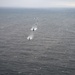 An aerial photograph shows a coordinated naval or maritime formation over open ocean under overcast skies. Multiple vessels are arranged in a column formation moving away from the camera, flanked by what appear to be aircraft flying at low altitude on either side. The scene suggests a military or coast guard exercise or escort operation.